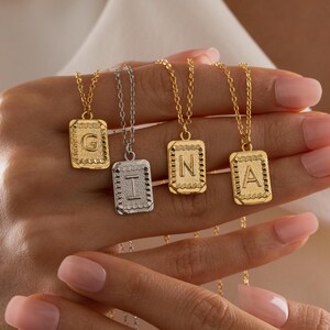 May include: A selection of initial pendant necklaces in gold and silver. Each necklace has a rectangular pendant with a letter. The pendants hang from fine gold chains. The necklaces are shown against a white backdrop.