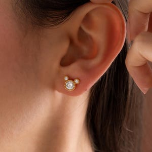 May include: Gold-colored stud earrings in a Mickey Mouse design. The center of the earring features a round, clear gemstone surrounded by smaller stones. The two smaller circles are also adorned with gemstones.