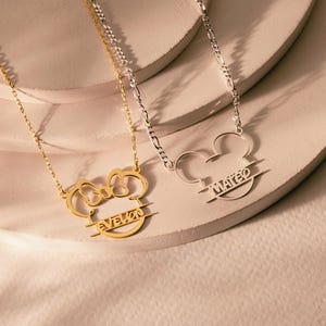 Peut inclure: Deux colliers avec un design de t&ecirc;te de Mickey Mouse. L'un est dor&eacute; avec le nom "Evelyn" et l'autre est argent&eacute; avec le nom "Mateo". Les deux colliers sont sur des cha&icirc;nes d&eacute;licates.