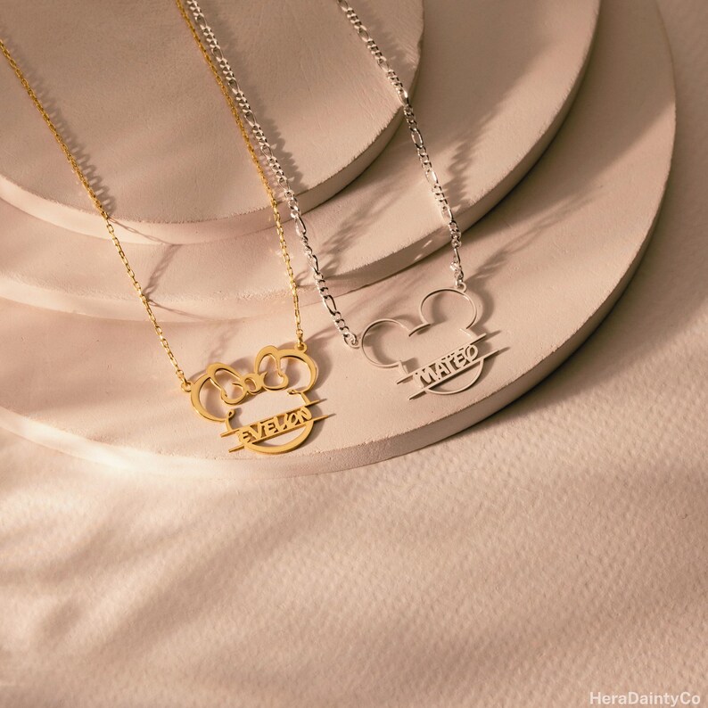 Peut inclure: Deux colliers avec un design de t&ecirc;te de Mickey Mouse. L'un est dor&eacute; avec le nom "Evelyn" et l'autre est argent&eacute; avec le nom "Mateo". Les deux colliers sont sur des cha&icirc;nes d&eacute;licates.