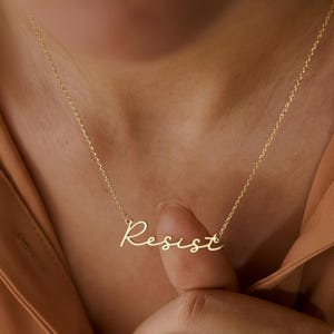 Op de afbeelding: Een delicate goudkleurige ketting met het woord "Resist" in cursief schrift. De ketting wordt om de nek gedragen, met de hanger op de borst. De ketting is dun en de hanger heeft een handschriftstijl.