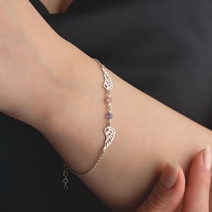 May include: A silver bracelet featuring wing-shaped charms and three colorful gemstones. The bracelet is worn on a wrist, showcasing its delicate design and intricate details. The gemstones are pink and purple.
