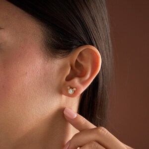 May include: A gold stud earring featuring a central round clear stone surrounded by a beaded metal border, with two smaller clear stones positioned at the top to create a rounded silhouette, secured on a gold post with a butterfly backing.