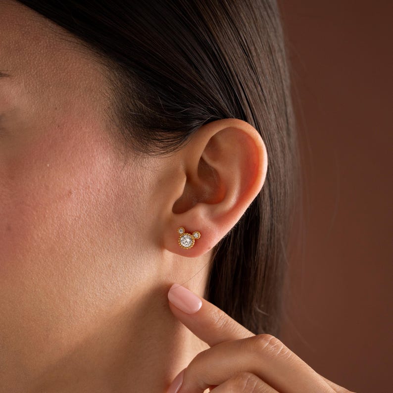 May include: A gold stud earring featuring a central round clear stone surrounded by a beaded metal border, with two smaller clear stones positioned at the top to create a rounded silhouette, secured on a gold post with a butterfly backing.