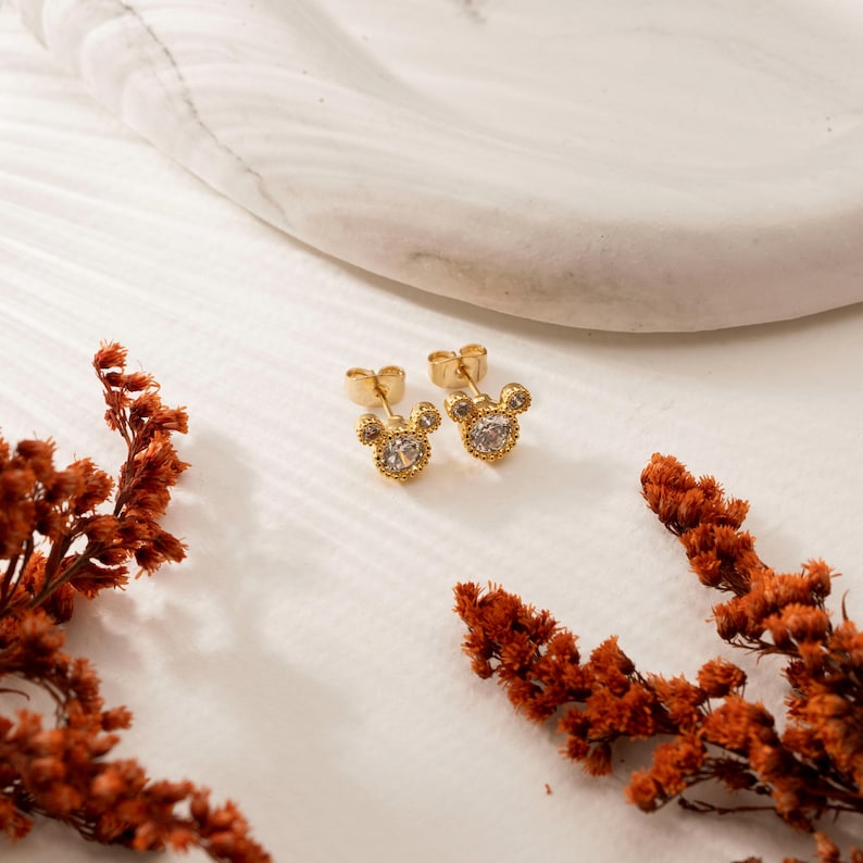 May include: A pair of gold stud earrings shaped like a mouse head silhouette. Each earring features a central round clear gemstone surrounded by two smaller circular accents at the top, creating a distinct three-circle design in a polished gold finish.
