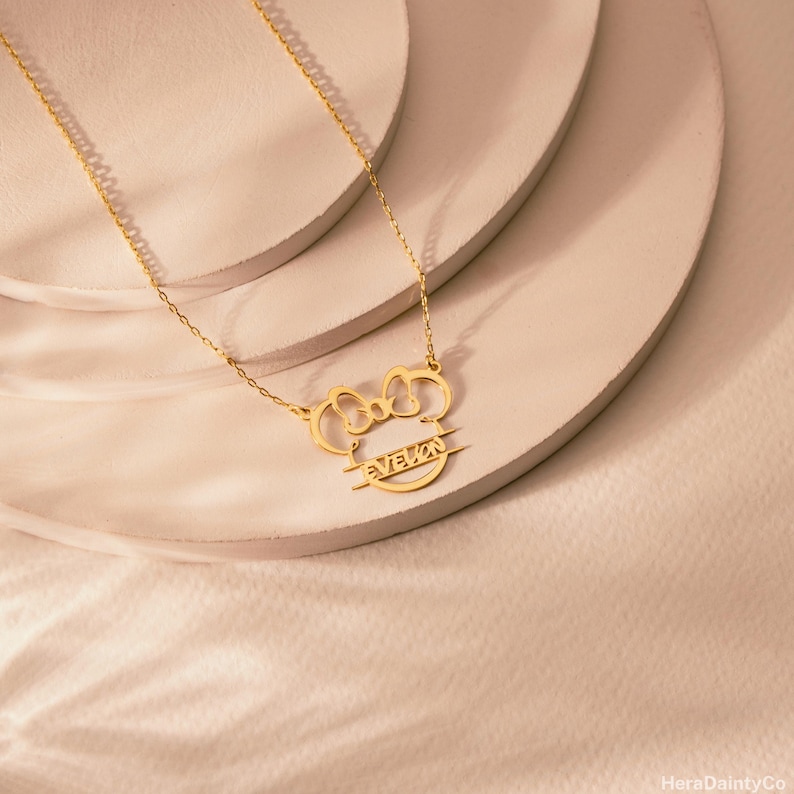 Peut inclure: Un collier dor&eacute; avec un pendentif en forme de n&oelig;ud et d'oreilles de souris. Le pendentif porte le nom "EVELYN". Le collier est pr&eacute;sent&eacute; sur une surface de couleur claire.