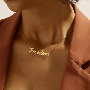 May include: A gold-coloured necklace spelling "Freedom" in a cursive font. The necklace is worn against a person's skin, with a rust-coloured blazer in the background.