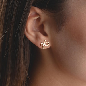 May include: A gold-coloured stud earring in the shape of a cursive letter "J" and "B" intertwined. The earring is worn on a person's ear, with the ear and part of the person's face visible in the image.
