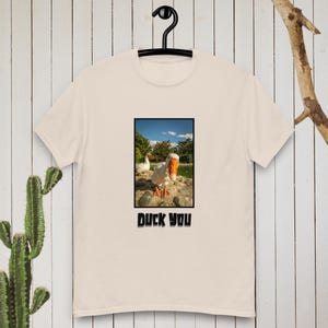 Könnte beinhalten: Cremefarbenes T-Shirt mit einem gerahmten Bild einer Gans mit orangefarbenem Schnabel und Füßen. Das Bild ist vor dem Hintergrund eines blauen Himmels und Felsen platziert. Der Text "DUCK YOU" ist unter dem Bild gedruckt.