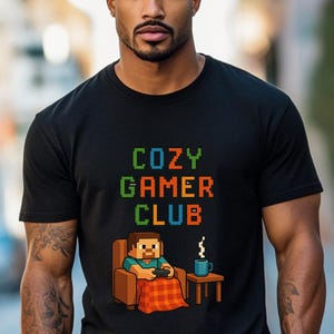 Può includere: T-shirt nera con la scritta "COZY GAMER CLUB" in lettere pixelate. Sotto, un'illustrazione pixelata di una persona che gioca a un videogioco, seduta su una sedia, con una tazza di caffè su un tavolino.