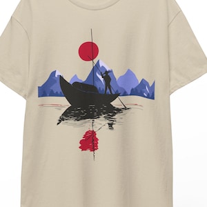 Può includere: T-shirt beige con un design a tema barca. Il design presenta un sole rosso, montagne e una persona su una barca. La barca si riflette nell'acqua. Il design utilizza nero, blu e rosso.