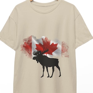 Può includere: T-shirt beige con un design della bandiera canadese, con una foglia d'acero rossa e una silhouette di alce nera. La bandiera ha accenti in stile acquerello rosso e bianco. La maglietta è appesa a una gruccia di legno.