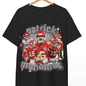 Può includere: T-shirt nero con grafica di un giocatore di football americano in uniforme rossa e la scritta "patrick mahomes". La grafica ha un aspetto vintage e usurato. La maglietta è appesa a una gruccia di legno.