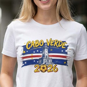 Può includere: T-shirt bianca con un design grafico con la scritta "CABO VERDE 2026". Il design comprende un faro, stelle e i colori della bandiera di Capo Verde. La maglietta è a maniche corte.