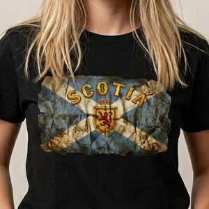 Può includere: T-shirt nera con un design invecchiato della bandiera scozzese. La bandiera presenta blu e bianco, con la scritta dorata "SCOTIA" e un emblema del leone rampante rosso. La t-shirt è a girocollo.