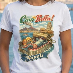 Può includere: T-shirt bianco con grafica colorata con la scritta "Ciao Bella!" e "Napoli." La grafica raffigura una scatoletta di sardine con un pesce all'interno, insieme alla Torre di Pisa e al Vesuvio. La maglietta è indossata da una persona.