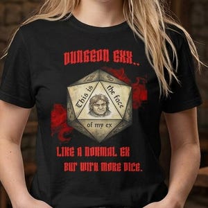 Può includere: Una t-shirt nera con la grafica di un dado a venti facce che mostra un volto al centro. Il testo recita: DUNGEON EXX.. This is the face of my ex LIKE A NORMAL EX BUT WITH MORE DICE. Il design presenta degli accenti di colore rosso.