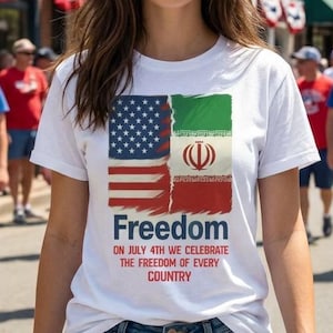 Può includere: Una maglietta bianca con una grafica raffigurante le bandiere americana e iraniana, con la parola "Freedom" e il testo "On July 4th we celebrate the freedom of every country." La maglietta è indossata da una persona tra la folla.