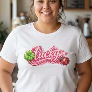 Può includere: Una maglietta bianca con la scritta "Lucky" in corsivo rosa, accompagnata da un quadrifoglio verde e una coccinella rossa. La maglietta è indossata da una persona in un bar.