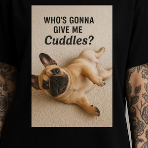 Pode incluir: Camiseta preta com um buldogue francês castanho claro deitado de costas. O texto "WHO'S GONNA GIVE ME CUDDLES?" está acima do cão. A camisa é de gola redonda.