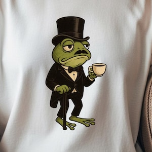 Puede incluir: Camiseta blanca con un dibujo de una rana con esmoquin negro, sombrero de copa y taza de té. La rana tiene bigote y bastón. El diseño es una representación humorística de un personaje sofisticado.