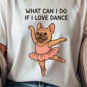 Pode incluir: Camiseta branca com o texto "WHAT CAN I DO IF I LOVE DANCE". A camiseta apresenta uma ilustração de desenho animado de um buldogue francês usando um tutu rosa e sapatilhas de balé, em uma pose de dança.