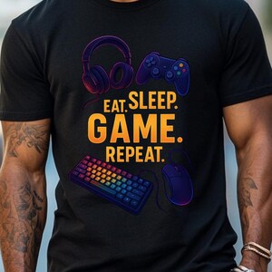 Può includere: T-shirt nera con la scritta "EAT. SLEEP. GAME. REPEAT." e grafica a tema gaming. Il design include cuffie, un controller di gioco, una tastiera e un mouse. Il testo è arancione e giallo.