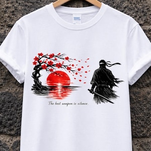 Può includere: T-shirt bianca con grafica nera e rossa di un ninja con una spada, un albero con fiori rossi e un sole rosso sull'acqua. Il testo "The best weapon is silence" è stampato sotto.