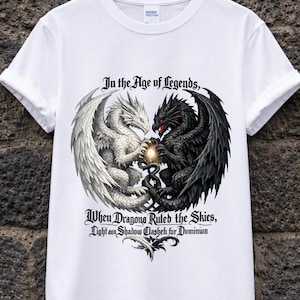 Può includere: T-shirt bianca con una grafica di due draghi, uno bianco e uno nero, uno di fronte all'altro. Il testo "In the Age of Legends, When Dragons Ruled the Skies, Light and Shadow Clash for Dominion" è stampato sopra e sotto i draghi.