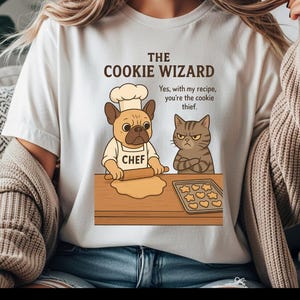 Könnte beinhalten: Weißes T-Shirt mit einer Cartoon-Illustration eines Hundes und einer Katze, die Kekse backen. Der Hund, der eine Kochmütze und eine Schürze trägt, rollt den Teig aus. Die Katze sieht verärgert aus. Text "The Cookie Wizard".
