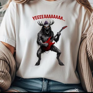 Può includere: T-shirt bianca con la grafica di un mostro che suona una chitarra elettrica rossa. Il mostro ha le corna ed è grigio scuro con i capelli viola. Il testo "YEEEEAAAAAAA..." è sopra il mostro.
