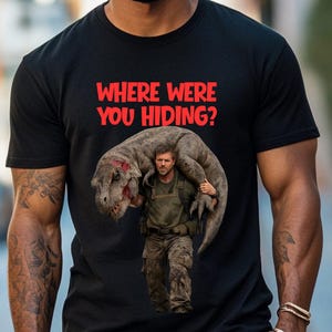 Puede incluir: Camiseta negra con el texto rojo "WHERE WERE YOU HIDING?" y un gráfico de un hombre cargando un dinosaurio. El hombre lleva pantalones de camuflaje y una camisa verde. El dinosaurio es gris y marrón.