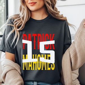 Può includere: T-shirt grigio scuro con il nome "PATRICK" in rosso, il numero "15" in bianco e il nome "MAHOMES" in giallo. La maglietta è in stile casual.