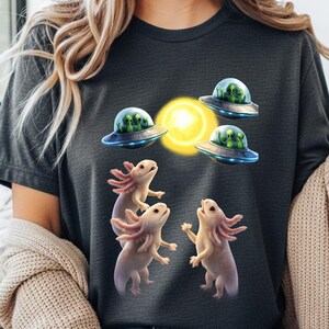 May include: Dark gray t-shirt featuring a whimsical design of axolotls and UFOs. The graphic includes three axolotls with pink gills and a yellow sun, with three UFOs each containing green aliens. A fun and unique design.