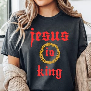 Può includere: T-shirt grigio scuro con la scritta "Jesus is King" in rosso, con caratteri gotici. La parola "is" è all'interno di una corona di spine dorata. Un capo d'abbigliamento casual.