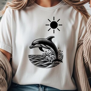 Könnte beinhalten: Weißes T-Shirt mit einer schwarz-weißen Grafik eines aus dem Meer springenden Delfins, darüber eine Sonne. Das Design ist eine detaillierte Illustration, ideal für Meeresliebhaber.