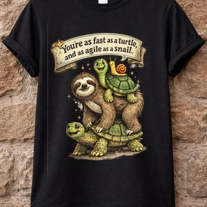 May include: Black t-shirt featuring a whimsical illustration of a sloth, two turtles, and a snail. The sloth is carrying the turtles, and the snail is on top. A banner above reads, "You're as fast as a turtle, and as agile as a snail."