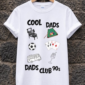 Può includere: T-shirt bianca con la scritta "COOL DADS" e "DADS CLUB 90s". La maglietta presenta illustrazioni in bianco e nero di un flipper, un tavolo da biliardo, un pallone da calcio, carte da gioco, un furgone e un cocktail.