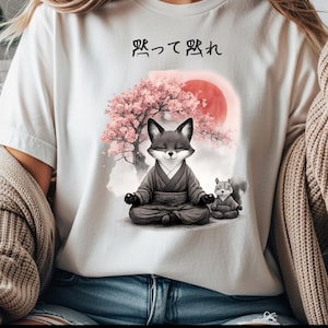Può includere: T-shirt bianca con un'illustrazione serena di una volpe e uno scoiattolo che meditano in abiti tradizionali, sullo sfondo di un albero di fiori di ciliegio e un sole rosso. Testo giapponese sopra.
