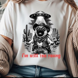 Può includere: T-shirt bianca con grafica di un carlino con cappello e gilet da cowboy, rapito da un UFO. Il testo rosso "I'm still too young" è stampato. Il design è ambientato su uno sfondo desertico.