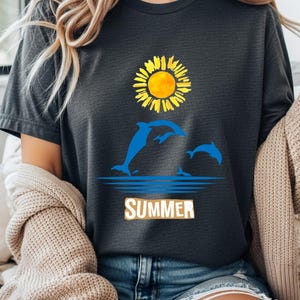 Könnte beinhalten: Dunkelgraues T-Shirt mit einer gelben Sonnenblume und blauen Delfinen, die aus dem Wasser springen. Das Wort "SUMMER" ist in Weiß und Beige gedruckt. Das Shirt ist ein lässiger Stil mit kurzen Ärmeln.