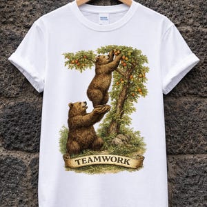 May include: White t-shirt featuring a detailed illustration of two brown bears. One bear is reaching for apples in a tree, while the other bear is standing below. The word "TEAMWORK" is on a banner.