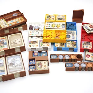 Può includere: Una collezione di organizzatori di giochi marroni, rossi, gialli, blu e bianchi pieni di carte, gettoni e componenti di gioco. Gli organizzatori sono progettati per contenere vari componenti di un gioco da tavolo, con carte che presentano illustrazioni di animali e paesaggi.