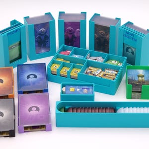 Può includere: Una collezione di organizzatori di giochi e porta carte color turchese. L'immagine mostra vari contenitori, tra cui scatole con le scritte "PROMO" e "NOVI SISTEN", e diversi mazzi di carte con disegni colorati. Gli organizzatori sono progettati per contenere i componenti del gioco.