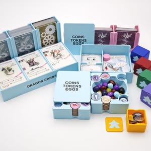 Könnte beinhalten: Ein hellblauer Spiel-Organizer mit Fächern für Karten, Spielsteine und Würfel. Der Organizer enthält Boxen mit der Aufschrift "Dragon Cards" und "Coins Tokens Eggs". Verschiedene Spielteile, darunter farbige Würfel und Spielsteine, sind sichtbar.