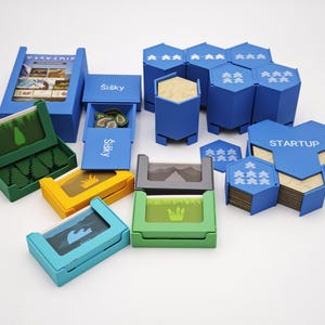 May include: A collection of board game components in various colors. The set includes blue, green, yellow, and gray boxes, along with hexagonal pieces and cards. Some boxes have text, including "STARTUP" and "Šišky". The game pieces are arranged on a white surface.