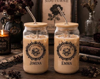 May include: Two clear glass tumblers with bamboo lids and metal straws, each filled with iced coffee. The glasses feature a black circular graphic with a skull, roses, and the text "JUST A LITTLE RAY OF" above the names "JIMENA" and "EMMA".