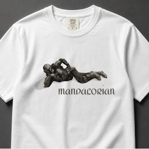 May include: White t-shirt featuring a detailed illustration of a character from the Mandalorian series. The character is depicted in a relaxed pose, with the word "Mandalorian" printed below in a stylized font. The shirt has a crew neck and short sleeves.