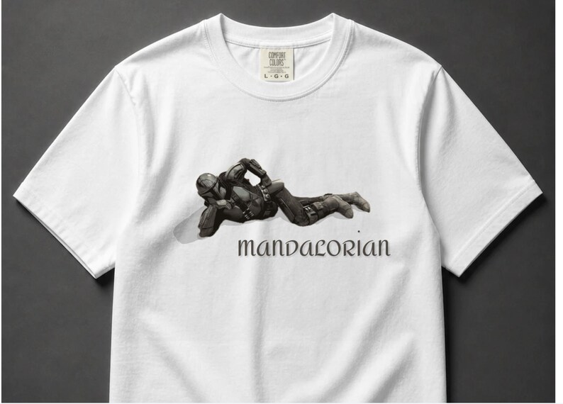 May include: White t-shirt featuring a detailed illustration of a character from the Mandalorian series. The character is depicted in a relaxed pose, with the word "Mandalorian" printed below in a stylized font. The shirt has a crew neck and short sleeves.