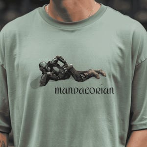 May include: Sage green t-shirt featuring a graphic of a character in black armor, lying down. The word "mandalorian" is printed in a stylized font below the image. The shirt has a crew neck and short sleeves.